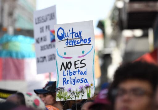 Foto de manifestación de personas LGBTQ+. Una persona levanta un cartel que dice quitar derechos no es libertad religiosa.